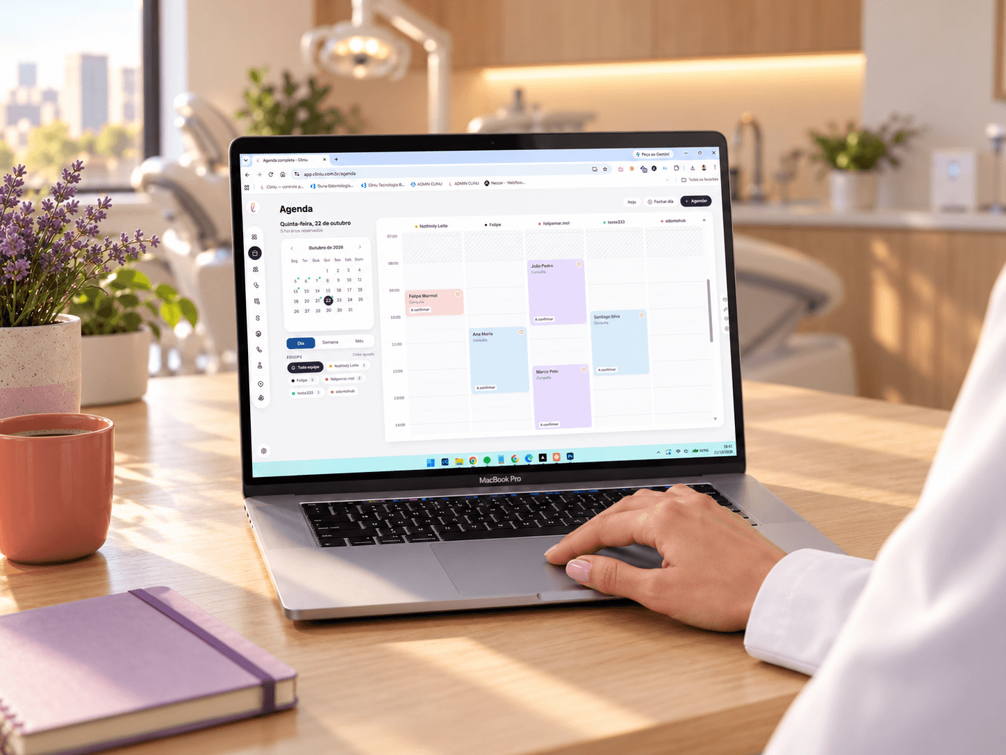 Notebook numa mesa de consultório com a agenda da Cliniu aberta, a mão de uma dentista de jaleco no touchpad